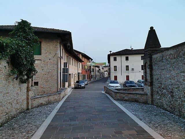 Scorcio di una suggestiva via cittadina medievale lastricata in pietra, fiancheggiata da antichi palazzi storici con balconi fioriti e piante rampicanti.